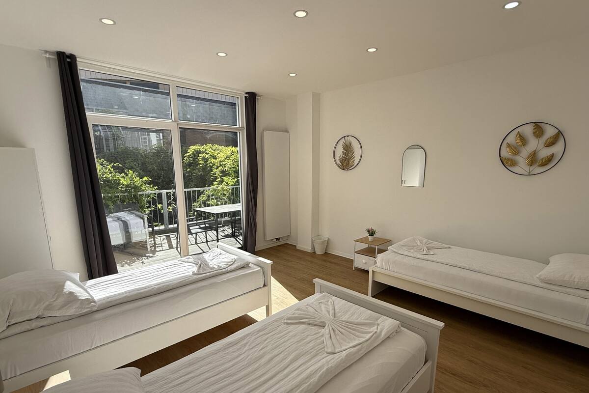 A hotel room with two single beds, a small table, a chair, a window with a view of greenery, and a wall-mounted mirror.