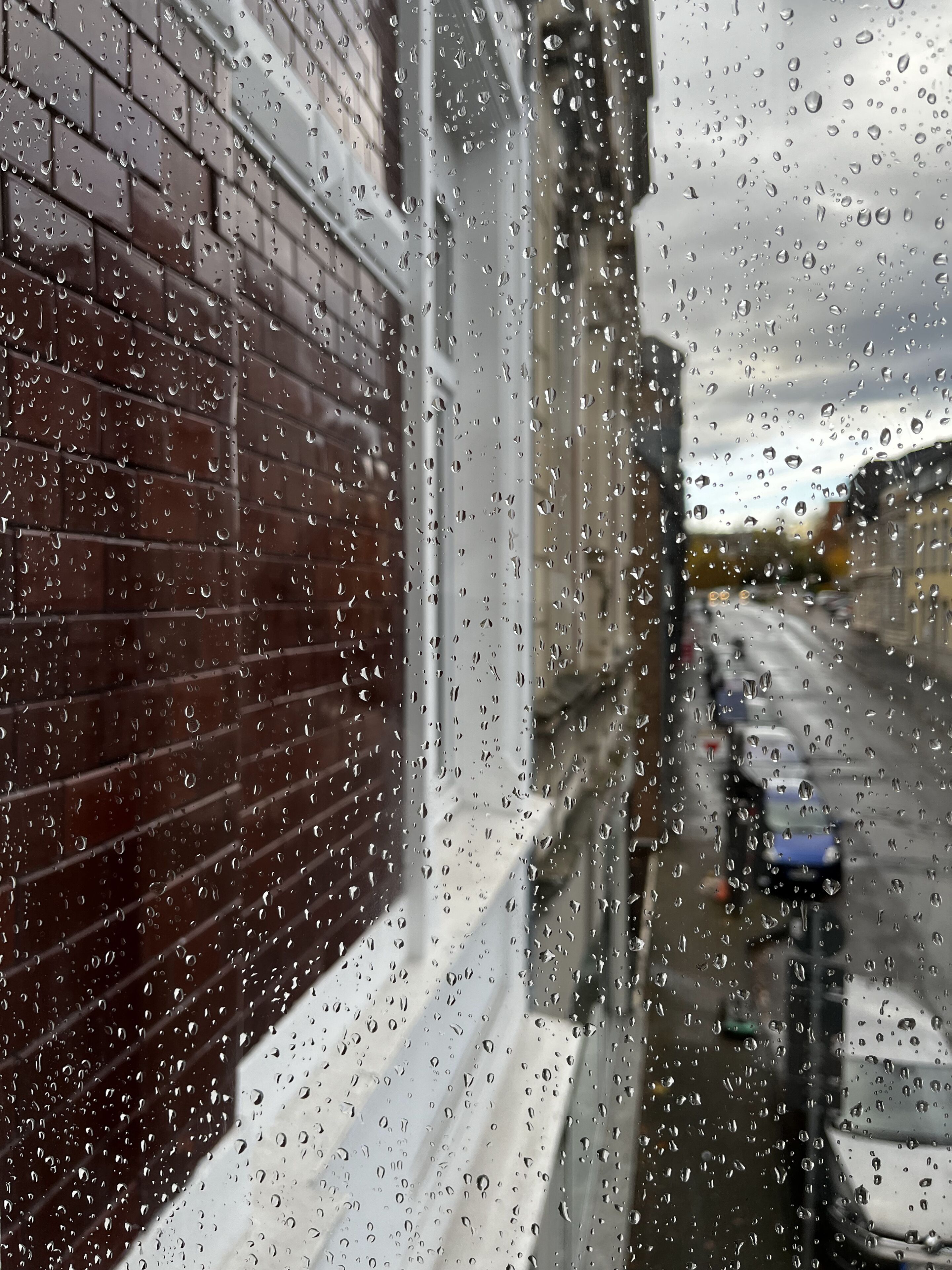 A window with raindrops, a brick wall, and a blurred street view.