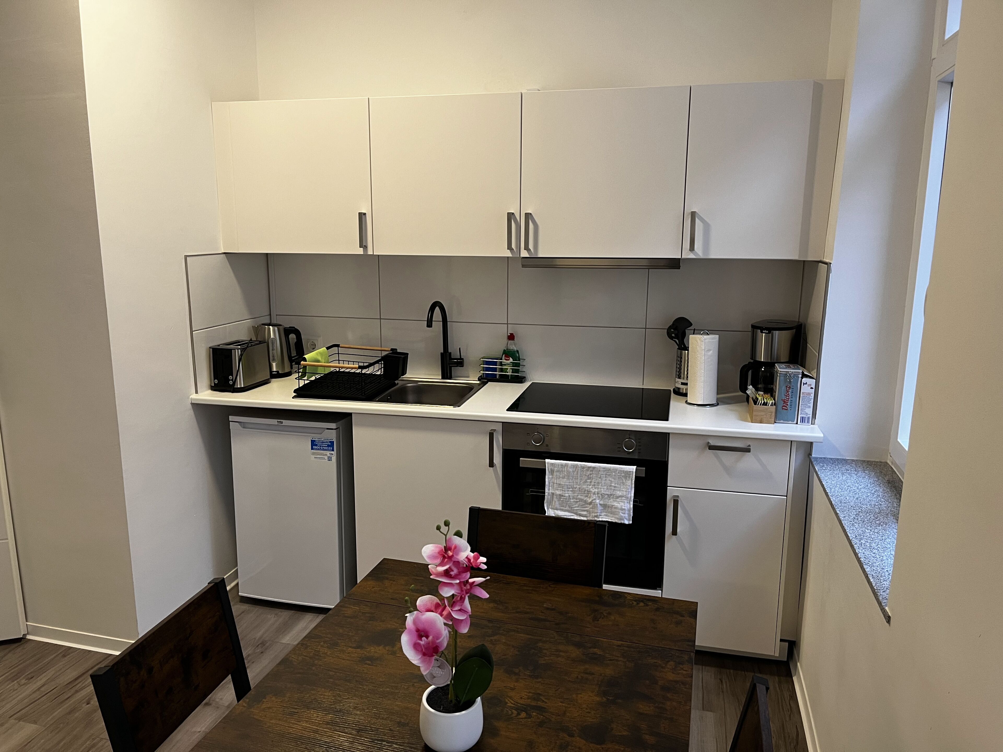 A compact kitchen with white cabinets, a sink, a small refrigerator, and a dining table with a potted orchid.