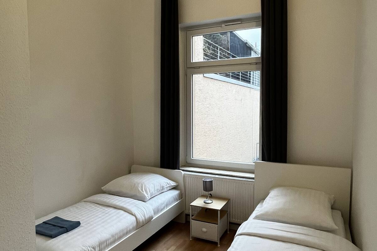 A small, single-room accommodation with two single beds, a window with curtains, a bedside table with a lamp, and a radiator.
