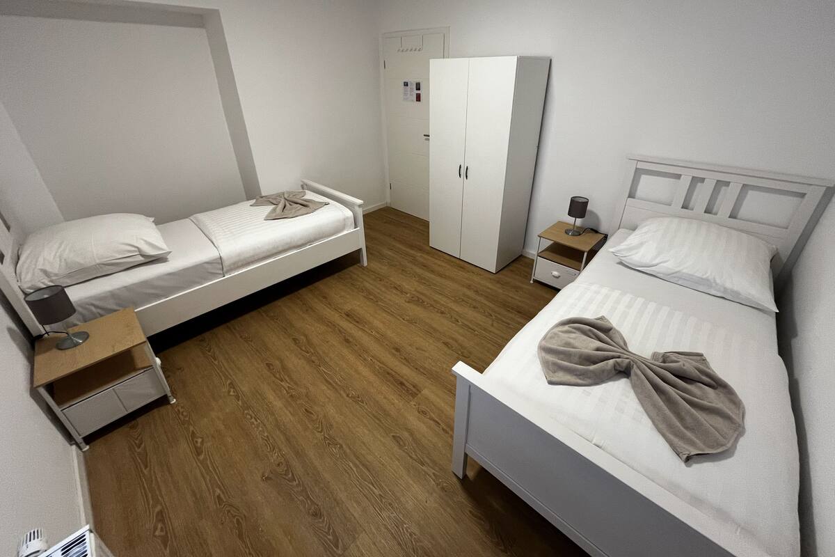 A bedroom with two single beds, a wardrobe, a bedside table with a lamp, and a small drawer unit.
