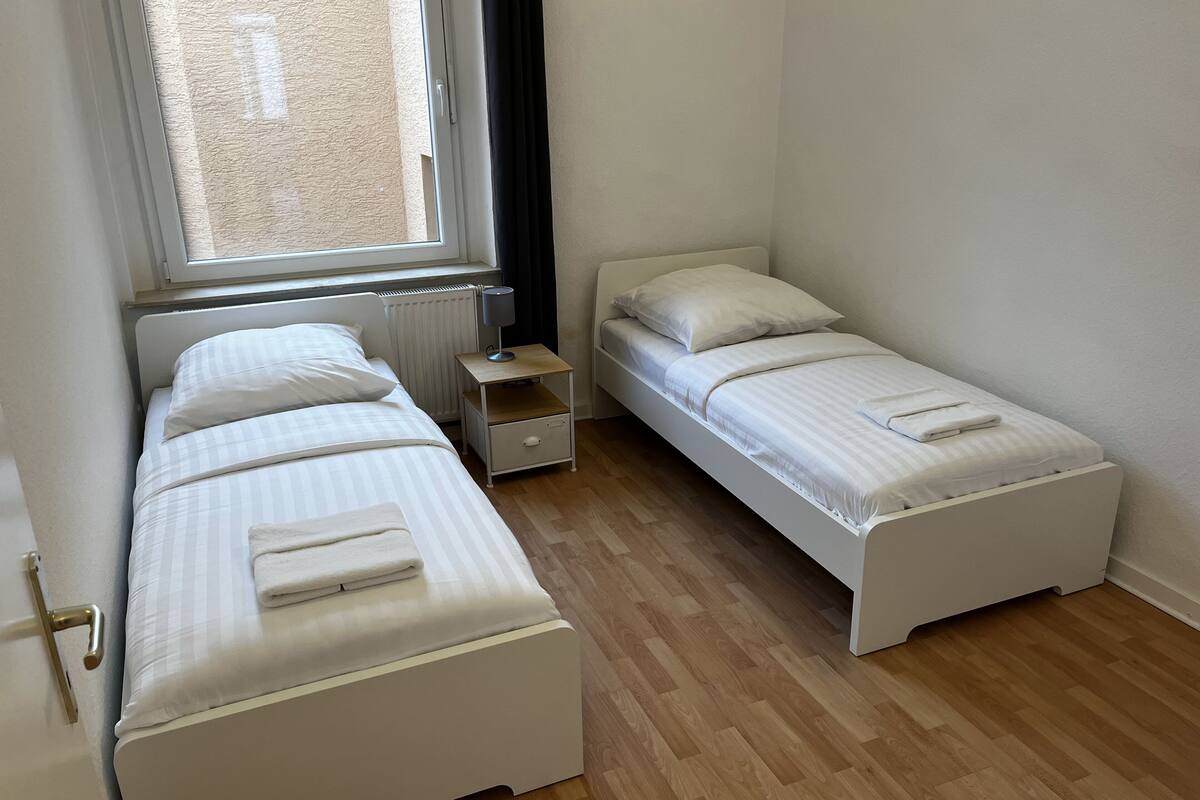 A room with two single beds, a small bedside table with a lamp, a radiator, and a window.