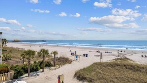 Ein Strand mit einer Holzsteganlage, die sich ins Meer erstreckt, ein Sandstrand mit Palmen und ein Gebäude mit einem Balkon mit Blick auf den Strand.