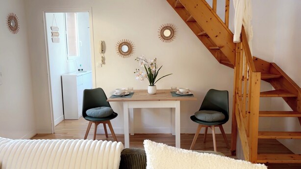 A dining area with a wooden staircase, two chairs, a table with a vase of flowers, and a kitchen in the background.