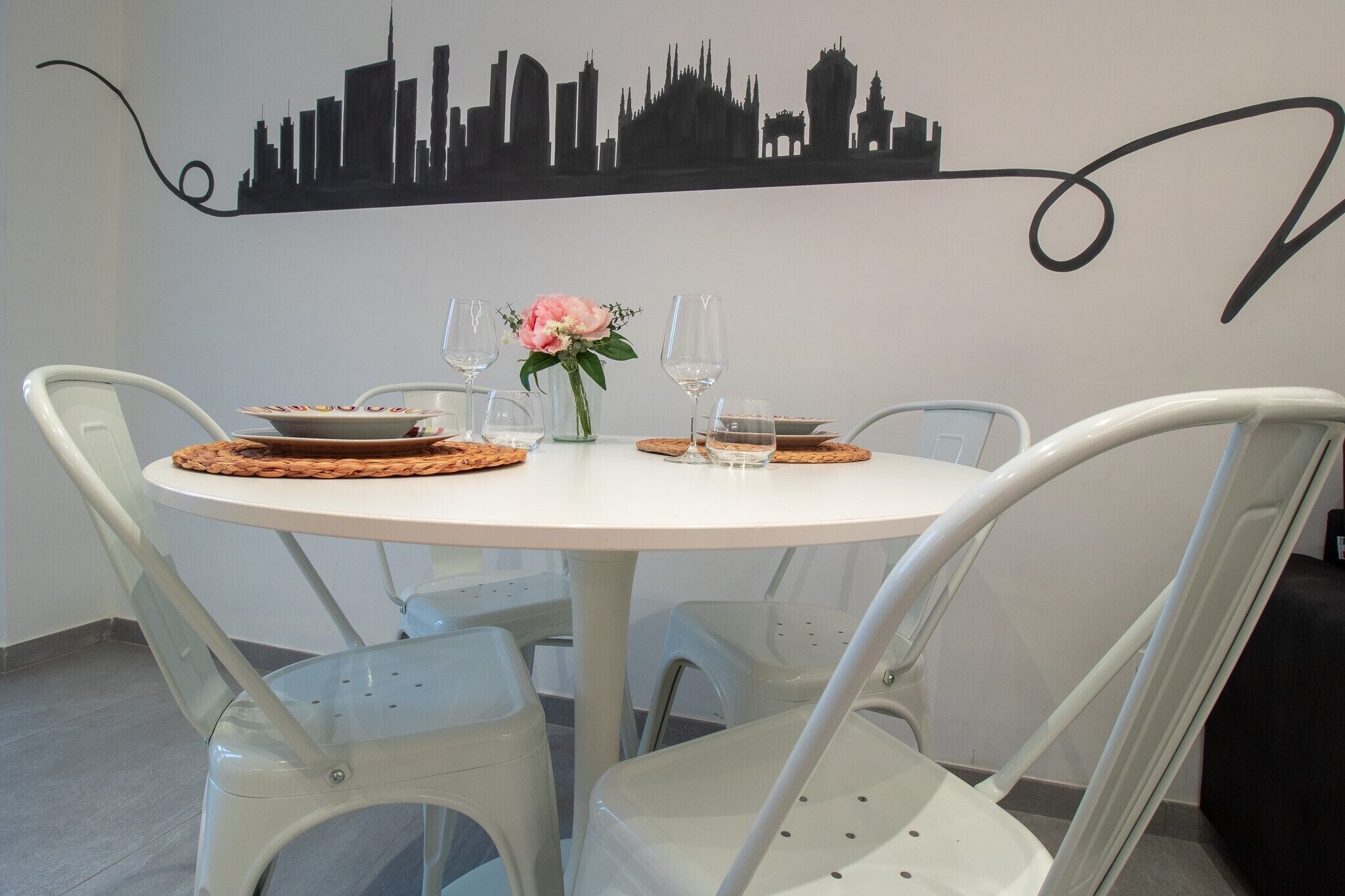 A round dining table with white chairs, a city skyline mural on the wall, and a vase with a pink flower.