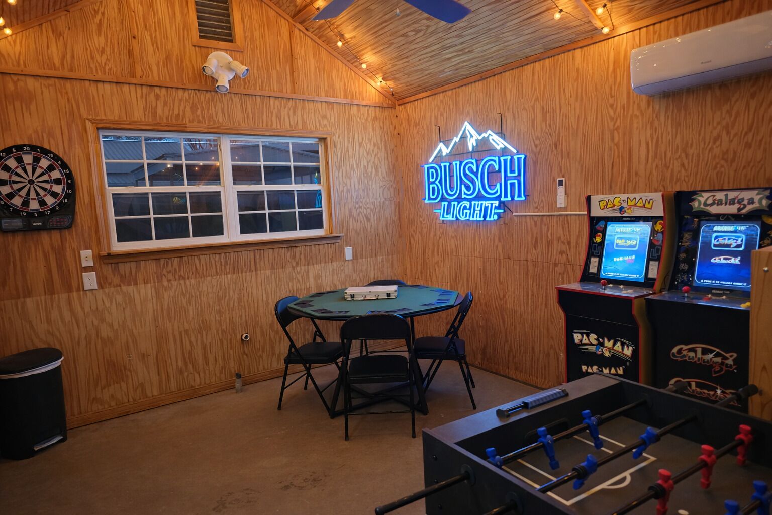 Een kamer met een dartbord, een neonbord waarop 'BUSCH LIGHT' staat, een tafelvoetbaltafel en speelautomaten.