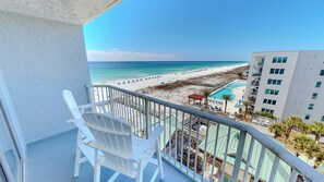Ein Balkon mit weißen Stühlen, Blick auf einen Strand, ein Schwimmbecken und ein Gebäude mit zahlreichen Fenstern.