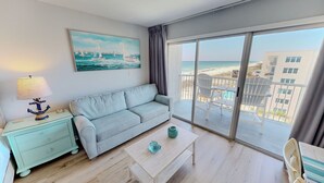 A living room with a sofa, a coffee table, a lamp, a painting, and a balcony with a view of the beach and buildings. - Top Floor Gulf Front Studio Suite IBR 705 w/Beach Service sleeps 6 with W/D (Fort Walton Beach)