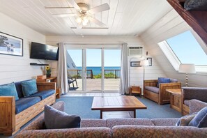 A room with a ceiling fan, a television, a sofa, a patterned couch, a wooden coffee table, and a view of the ocean through large sliding doors.