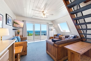 Ein gemütliches Zimmer mit Holzbalkendecke, Deckenventilator, Fernseher, Sofa, Tisch und einer Treppe.