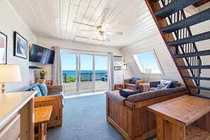 A room with a wooden ceiling, a ceiling fan, a TV mounted on the wall, a sofa, a table, and a staircase.