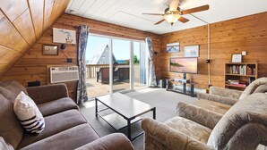 A cozy cabin interior with a wooden ceiling, a ceiling fan, a large sliding glass door to a deck, and a bookshelf. - Juniper Presents: Chalet #25 Walk to Beach Chalet & Resort Amenities (Stevensville)