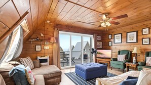 Ein gemütliches Blockhaushaus-Interieur mit Holzbalkendecke, einer Couch, einem blauen Hocker, einem hölzernen Bett und einem Balkon mit Aussicht.