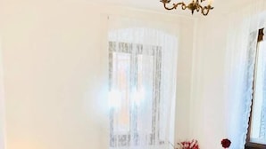 A neatly made bed with white and red pillows, a bedside table with a lamp, and a window with sheer curtains. - App. Accanto ai Mercatini di Natale (Trento)