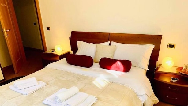 A neatly made bed with white linens and red pillows, a wooden headboard, and bedside tables with lamps. - App. Accanto ai Mercatini di Natale (Trento)
