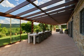 Una veranda esterna con una pergola, un tavolo lungo con sedie e un muro in pietra.