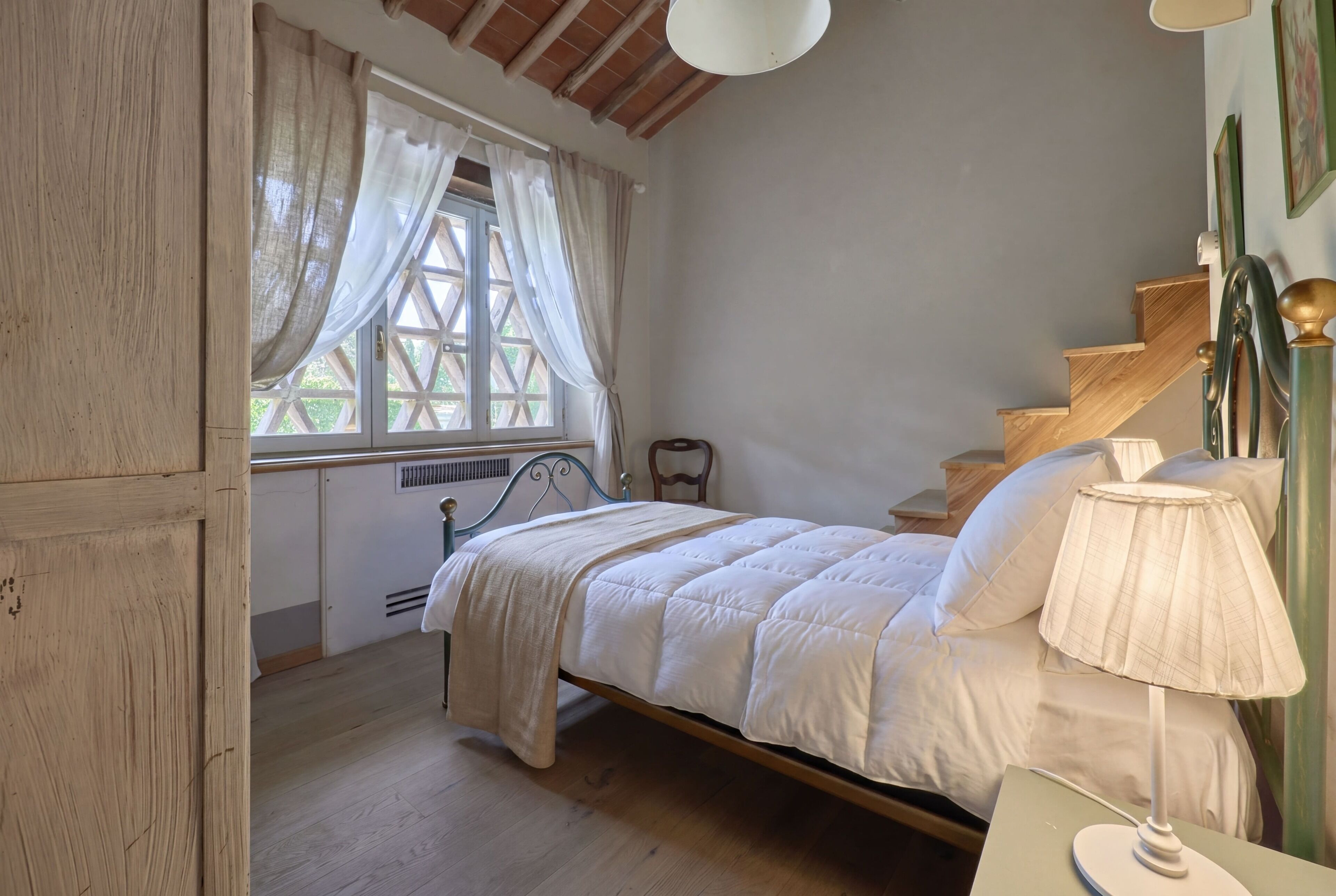 A bedroom with a wooden bed, a window with curtains, a nightstand with a lamp, and a staircase.