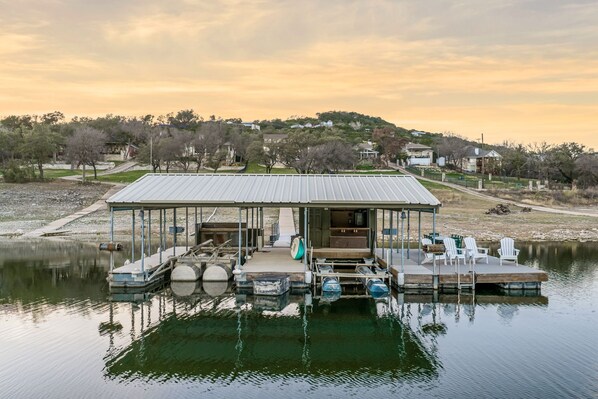 Un muelle techado con asientos y una mesa, rodeado de aguas tranquilas, con casas y árboles al fondo.