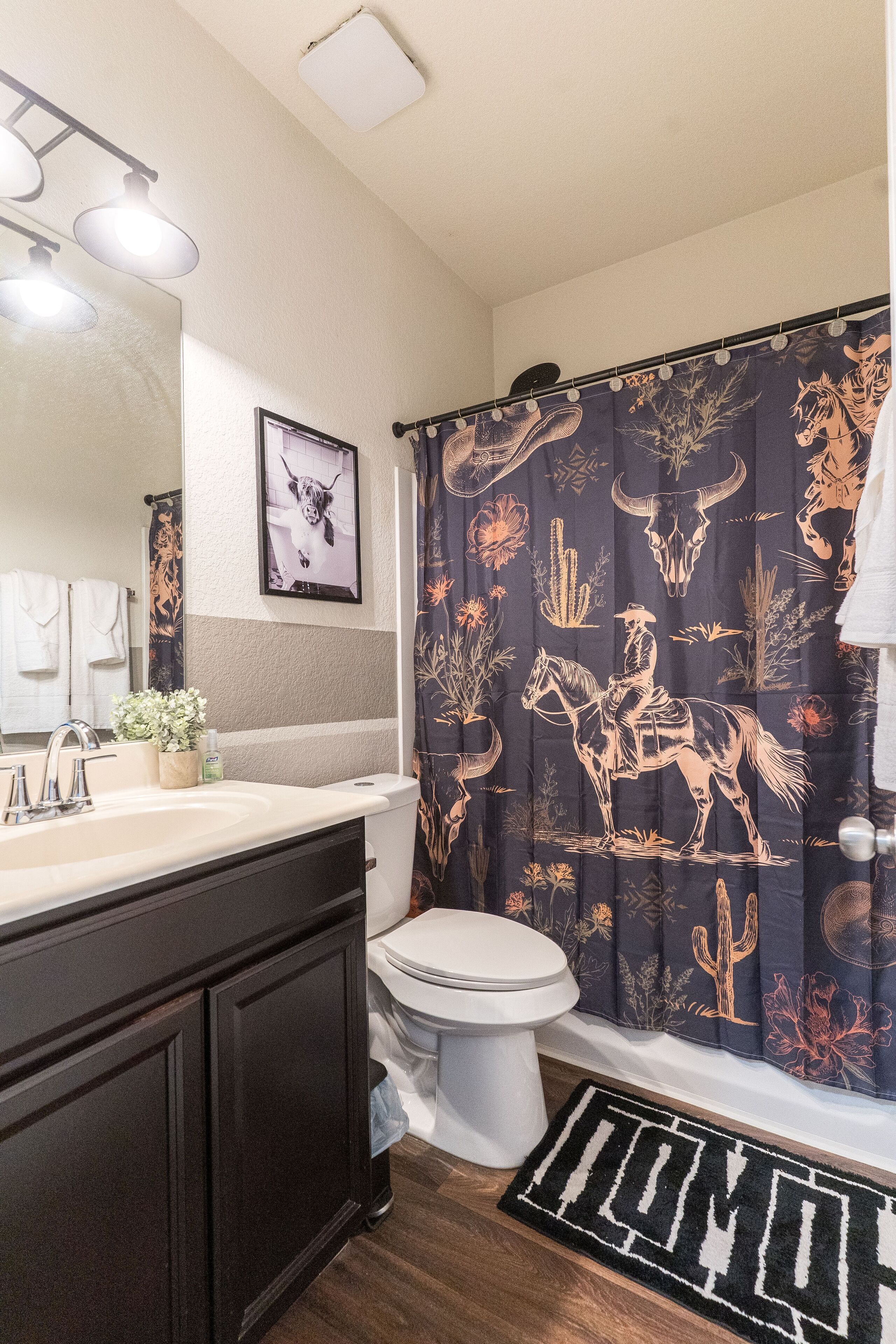 A bathroom with a dark shower curtain featuring a cowboy and cactus design, a white toilet, and a wooden floor.
