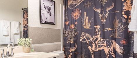 A bathroom with a dark shower curtain featuring a cowboy and cactus design, a white toilet, and a wooden floor.