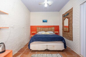 Une petite chambre à coucher accueillante, avec un mur en briques blanches, un lit simple recouvert d’une couverture bleue, une tête de lit en bois, une petite table avec une bouilloire et un ventilateur de plafond.