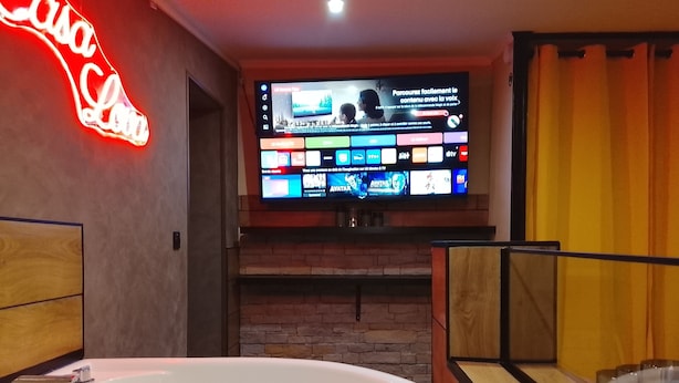 Een moderne badkamer met een groot bad, een televisie aan de muur die verschillende zenders toont, en een neonreclame.