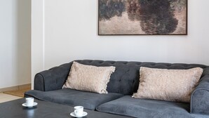Een grijze bank met twee beige kussens, een witte kop op een schotel, en een lijst met een schilderij van bomen aan de muur.