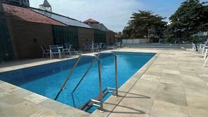 Una piscina con una escalera metálica, rodeada de sillas de descanso y un solárium con baldosas.