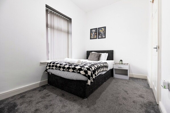 A bedroom with a bed, a nightstand, a window with blinds, and two framed pictures on the wall. - Spacious 4BR Home Near Alton Towers (Stoke-on-Trent)