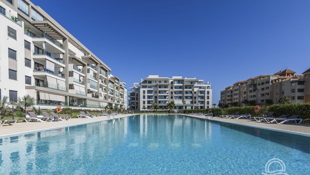 A swimming pool surrounded by modern buildings with balconies, clear skies, and lounge chairs along the poolside.