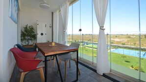 A balcony with a table and chairs, a potted plant, and a view of a sports field and pool.
