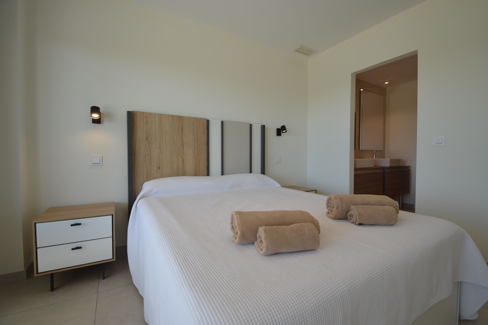 Un dormitorio moderno con una cama grande, dos mesitas de noche, un cabecero de madera y un baño visible a través de una puerta abierta.