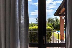 Blick durch ein Fenster mit Vorhängen auf einen Balkon mit Pool, Grünflächen und blauem Himmel.