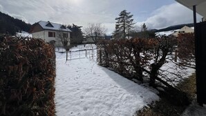 Een besneeuwde weg die naar een huis leidt, met een schoorsteen, omringd door bomen en een hek.