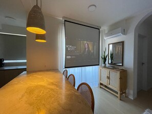 A modern dining area with a large table, chairs, a projector screen displaying a show, and a cabinet with plants. - Solarium Beach Club - Apartamento T1.304 (Itapoá)