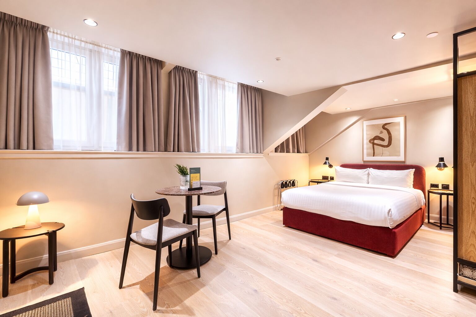 A modern hotel room with a bed, a desk, two chairs, a small table with a plant, and a painting on the wall.