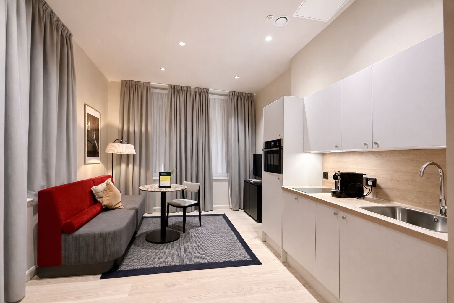 A modern hotel room with a sofa, a small table, a kitchen area with a sink, and a TV.
