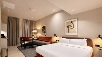 A hotel room with a bed, a desk, a chair, a sofa, a trash bin, and a lamp.