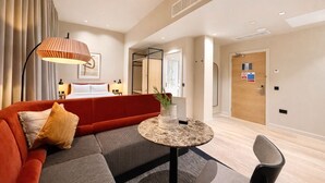 A modern hotel room with a red sofa, a round table with a vase, a bed with white linens, and a wooden door with a sign.