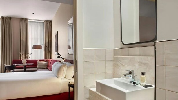 Habitación de hotel con una cama grande, un sofá rojo, una mesita, una lámpara y un baño con lavamanos y espejo.