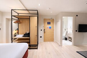 Ein Hotelzimmer mit Bett, einem Holzschrank, einem Schreibtisch mit Lampe und einem durchgegangenen Tür erkennbaren Badezimmer.