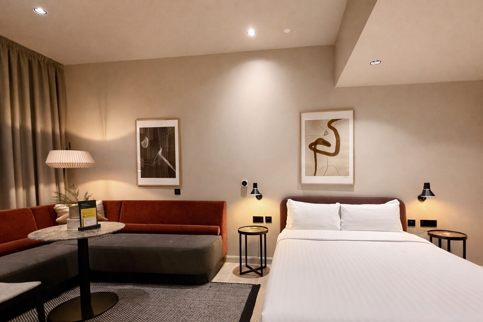 A hotel room with a bed, a sofa, a small table, two wall-mounted lamps, and two framed artworks.