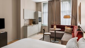 A modern hotel room with a large bed, a sofa, a small dining table, a kitchen area, and a large window with curtains.