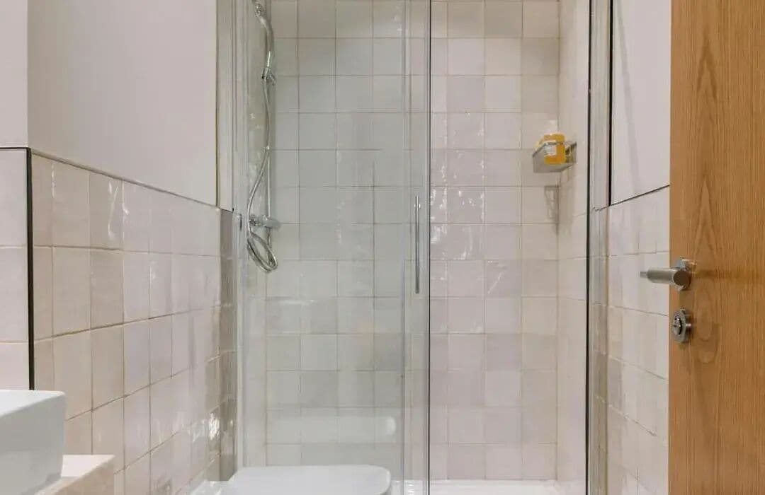 A shower with a glass door, a wooden door to the right, and a white tiled wall.