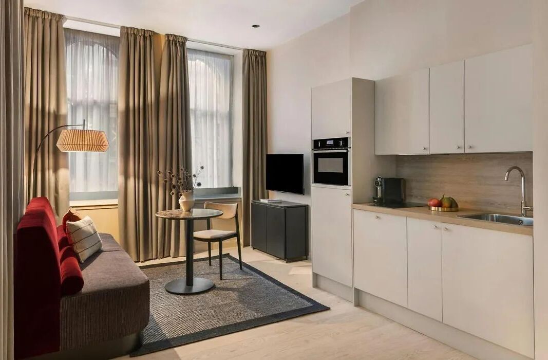 A modern living room with a sofa, a small round table, and a TV. There is a kitchen area with cabinets and a sink.