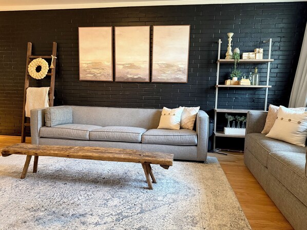 Un salon avec un canapé gris, un banc en bois, un mur en briques noires et une étagère ornée d’objets décoratifs.