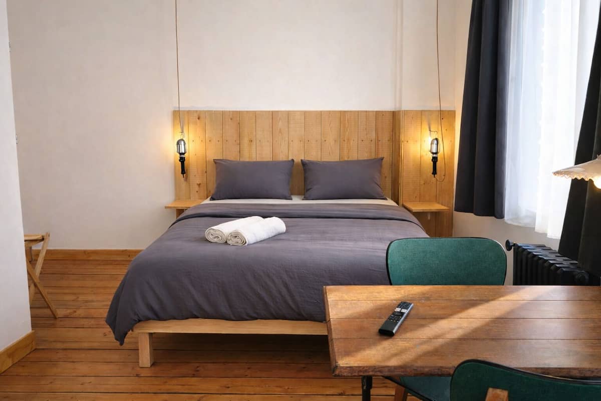 A hotel room with a wooden headboard, two bedside lamps, a bed with grey bedding, a rolled white towel, a wooden nightstand, a green chair, a wooden table, and a remote control.