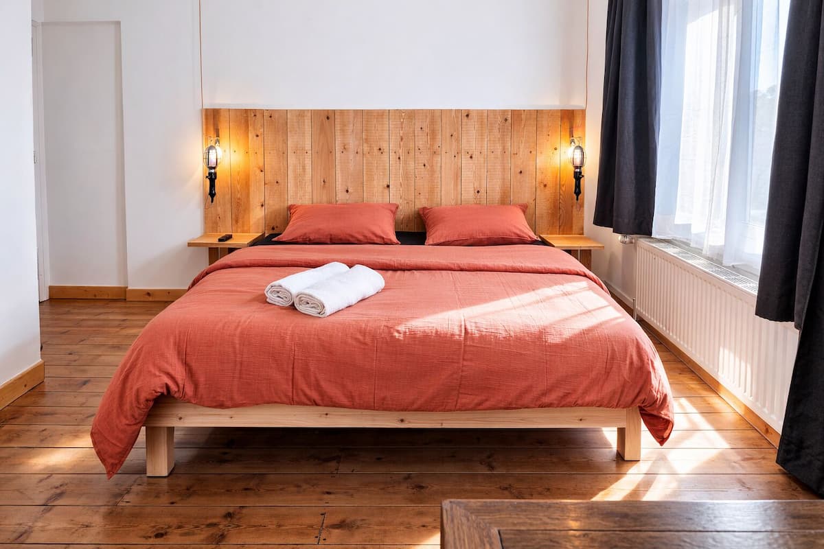 A neatly made bed with a red bedspread and two pillows, a wooden headboard, two bedside tables with lamps, and a window with curtains.