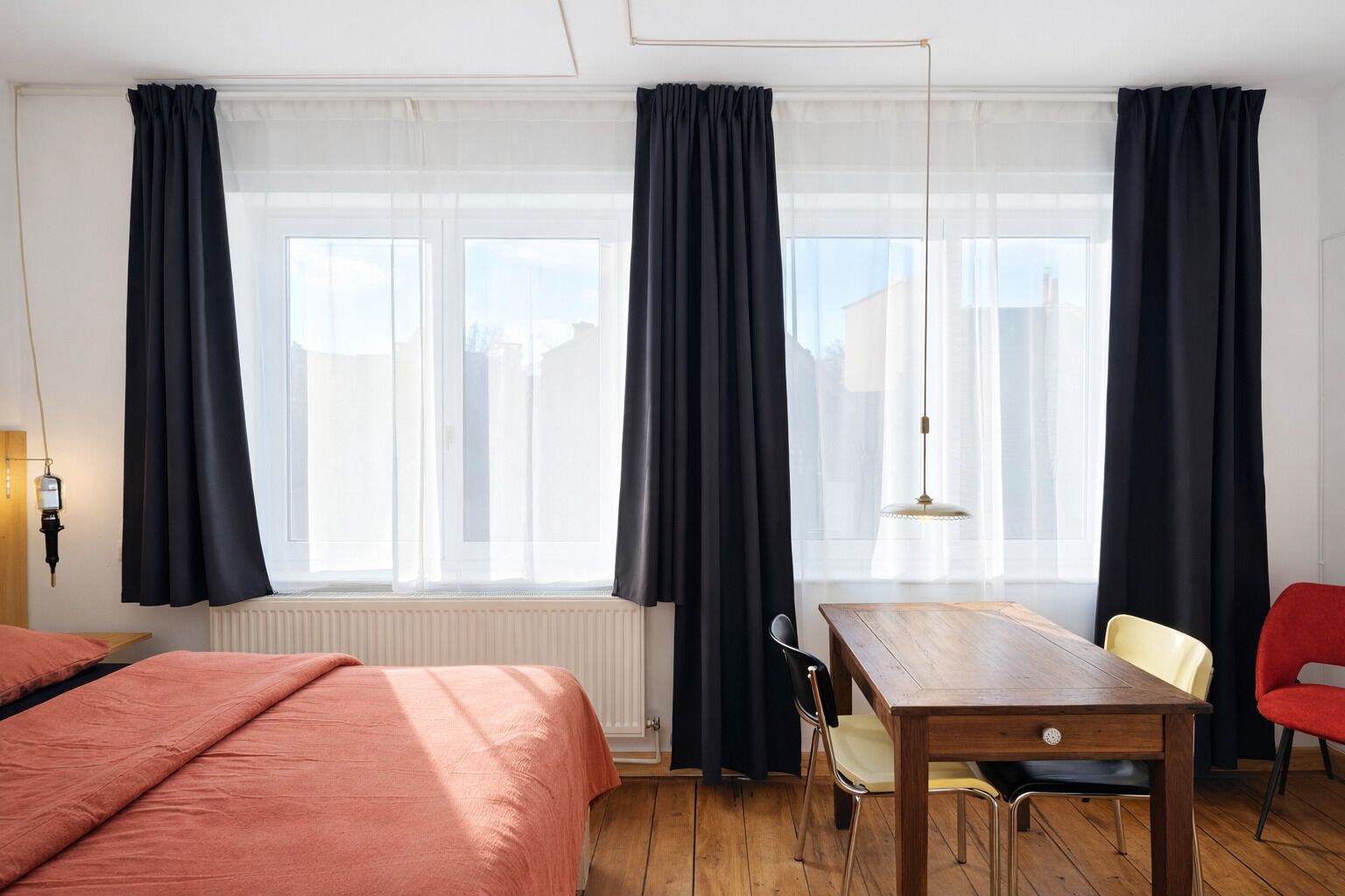 Une chambre d’hôtel avec un plancher en bois, un lit recouvert d’une courtepointe rose, une table en bois avec des chaises et une fenêtre avec des rideaux noirs.