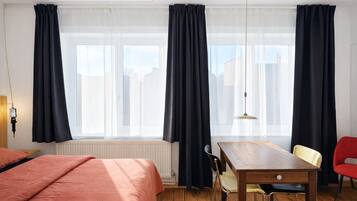 Une chambre d’hôtel avec un plancher en bois, un lit recouvert d’une courtepointe rose, une table en bois avec des chaises et une fenêtre avec des rideaux noirs.